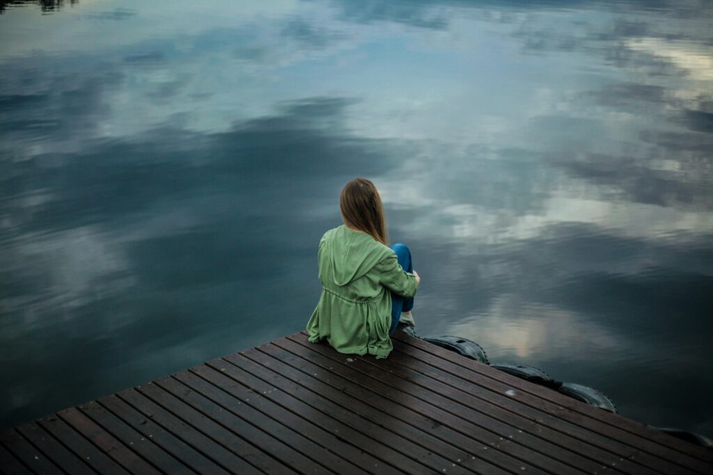 pexels-photo-2865901-2865901 A woman sits on a wooden dock, reflecting by a calm lake under a cloudy sky.